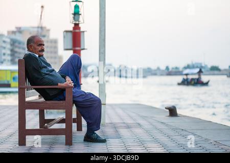 Älterer Mann, der sich auf Einer Holzbank am Ufer mit Einem Boot in der Ferne entspannt Stockfoto