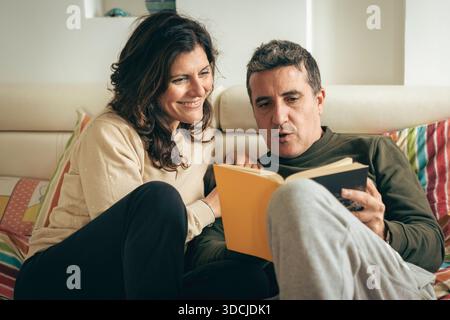 Paare mittleren Alters, die sich im Schlafzimmer entspannen. Ein Mann, der seiner lächelnden Frau ein Papierbuch vorliest, während er auf bunten Bettlaken liegt. Authentischer Lifestyle. Stockfoto