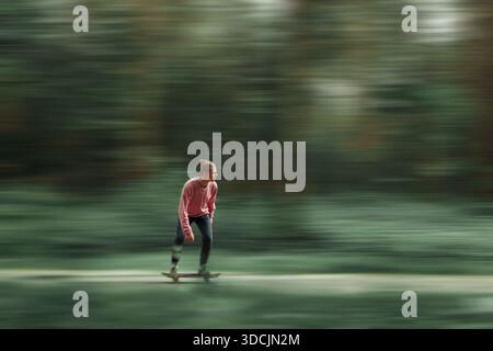 Junger Mann, der mit Bewegungsunschärfe auf einem Skateboard fährt, legere Kleidung trägt, Geschwindigkeit und Freiheit im Freien in einem grünen Waldhintergrund genießt. Dynamisch Stockfoto