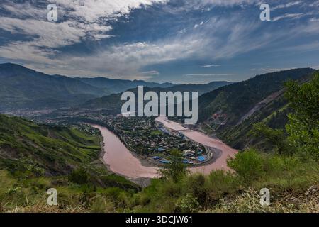 Blick auf den schlammigen Fluss, der sich durch üppige grüne Berge schlängelt, eine Stadt, die sich im Tal unter einem bewölkten Himmel befindet, Naran, Khyber Pakhtunkhwa, Pakistan. Stockfoto