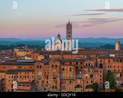 Aus der Vogelperspektive auf Terrakotta-Dächern, die unter einem weichen Dämmerungshimmel kaskadieren, dominiert vom Duomo di Siena und Torre del Mangia, Siena, Toskana, Italien. Stockfoto