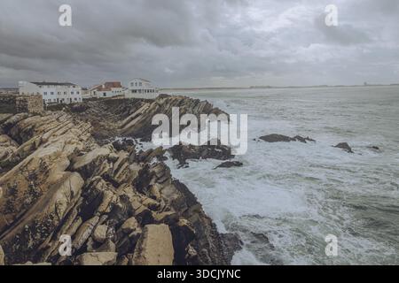 Blick auf zerklüftete Klippen treffen auf das aufgewühlte Meer unter einem dramatischen Himmel, mit weißen Gebäuden am Rand, Peniche, Bezirk Leiria, Portugal. Stockfoto