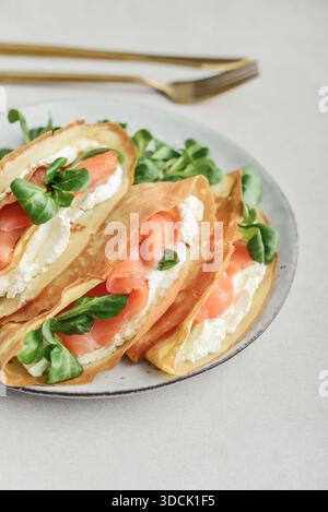 Crepes oder dünne Pfannkuchen mit leicht gesalzenem Lachs, Weichkäse und Maissalat oder Lammsalat auf einem Teller aus nächster Nähe auf leicht strukturiertem Backgro Stockfoto