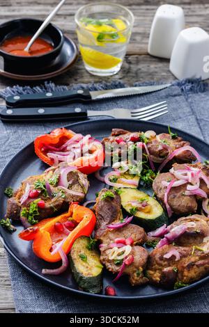 Gegrillter Schweinehals mit gebratener Zucchini, Paprika und roten Zwiebeln auf Teller. Serviert mit Tomatensauce, Wasser mit Zitrone und Besteck auf rustikalem Holz ta Stockfoto