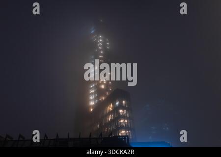 Filmische Aufnahme eines modernen Wolkenkratzers, der nachts in dickem Nebel verschwindet, mit leuchtenden Bürofenstern. Nebelige nächtliche Stadtlandschaft mit beleuchtetem o Stockfoto