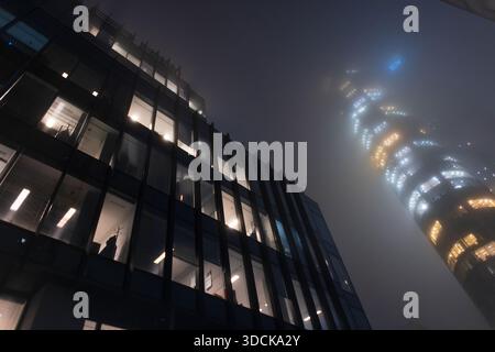 Moody-Architektur im Cyberpunk-Stil mit einem Hochhaus, das durch dichten Stadtnebel und Dunkelheit erstrahlt. Nebelige nächtliche Stadtlandschaft mit ausgeschalteter Beleuchtung Stockfoto