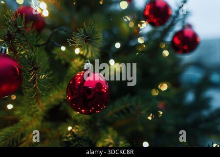 Nahaufnahme von roten Weihnachtskugeln, die an einer Kiefer mit Bokeh-Lichtern hängen. Schöne festliche, glänzende rote Ornamente auf üppig grünen Kiefernzweigen. Creati Stockfoto