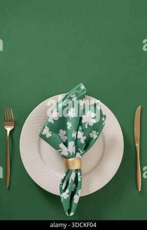 Festliche Tischdekoration am St. Patricks Day mit weißer Platte, grüner Schamrock Serviette auf grünem Hintergrund. Vertikales Format. Vorlage. Flach gelegen, Feiertagsfeier Stockfoto