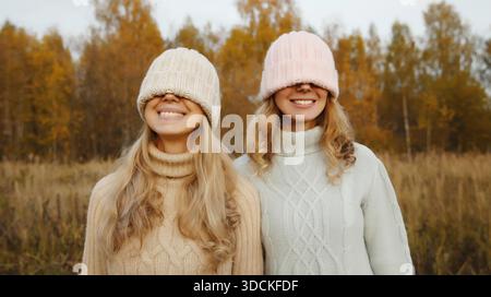 Porträt von zwei glücklichen Freundinnen, die Spaß haben, strickte Wintermütze über ihr Gesicht zu ziehen, die ihre Augen bedeckt, lustige Freundinnen, die draußen im Herbstpark zusammenkommen || Model veröffentlicht Stockfoto