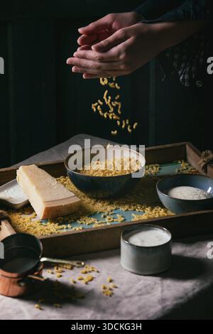 Junger Mann in Schwarz, der rohe ungekochte italienische Pasta-Coquillettes zum Kochen des Abendessens mit Parmesankäse, Sahne und Grießmehl auf Holztablett auf Küchen-Leinen-Tischtuch bestreut. Dunkler, rustikaler Stil. Stockfoto