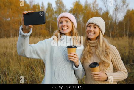 Glückliche zwei Freundinnen, die Selfie mit dem Telefon im Herbstpark machen, fröhliche Freundinnen lächeln und Spaß haben zusammen mit einer Tasse Kaffee im Freien, tragen Wintermütze, Pullover in der warmen Herbstsaison || Model veröffentlicht Stockfoto