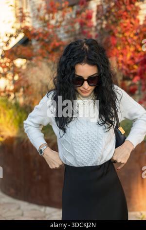 Herbstmodekonzept – elegante Frau im trendigen Outfit, die draußen mit herbstlichem Laubhintergrund posiert. Schönes Mädchen in einer modischen weißen Strickschnur Stockfoto