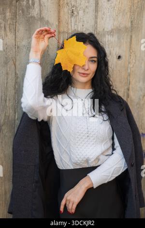 Fröhliche Frau mit lockigen Haaren, die ein gelbes Herbstblatt hält, einen weißen Pullover und einen grauen Mantel trägt, draußen im warmen Sonnenlicht vor einem rustikalen Lächeln lächelt Stockfoto