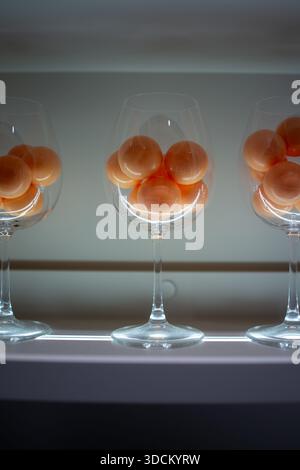 Drei Weingläser gefüllt mit orangefarbenen Kugeln in minimaler Stilllebenskomposition, sanftem Licht, sauberem Hintergrund und Symmetrie. Stockfoto