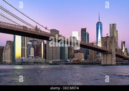 Die berühmte Brooklyn Bridge überspannt den East River und führt bei einem friedlichen, violetten Sonnenaufgang in die Skyline von Lower Manhattan. Stockfoto