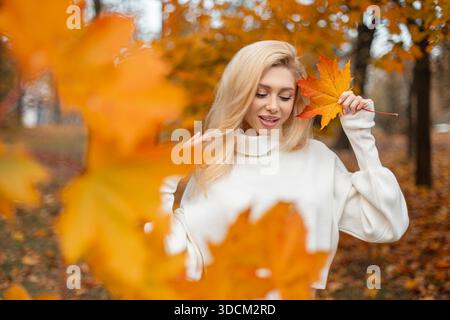 Fröhliches Mädchen, das ein Ahornblatt vor dem Gesicht hält, umgeben von Herbstfarben. Ein positives trendiges Mädchen hält ein leuchtend bunt gelbes Ahornblatt. Au Stockfoto