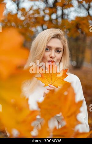 Glückliche Frau mit langen Haaren, die Ahornblatt vor dem Gesicht auf Herbsthintergrund hält. Ein fantastisches trendiges Mädchen küsst und hält einen bunten gelben Ahorn Stockfoto
