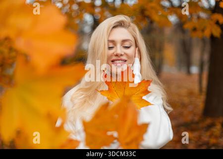 Attraktives Mädchen in weißem Outfit posiert im Herbstpark mit leuchtend orangefarbenen Blättern. Ein positives trendiges Mädchen hält ein leuchtend bunt gelbes Ahornblatt. Autum Stockfoto