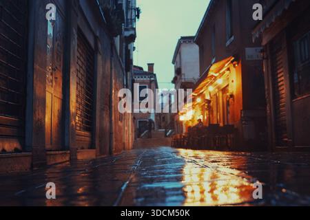 Reise italien venedig die feuchte Kopfsteinpflasterstraße, die warme Lichter reflektiert, sorgt für eine stimmungsvolle urbane Szene in der Abenddämmerung Stockfoto