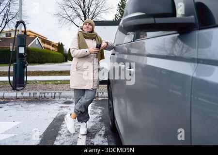 Eine ruhige Unabhängigkeit ist sichtbar, wenn eine Frau mittleren Alters neben ihrem Elektroauto wartet, während sie ihr Telefon überprüft und die Ladezeit in eine ruhige Persönlichkeit verwandelt Stockfoto