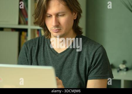 Ein junger Mann mit langen Haaren, der an seinem Schreibtisch sitzt und an einem Laptop arbeitet. Er schaut konzentriert auf den Monitor. Fernarbeit von zu Hause aus. Stockfoto