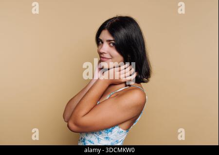 Eine Frau steht in einem Studio, ihre Hände auf ihren Haaren, während sie nachdenklich zur Seite schaut. Der Hintergrund ist eine helle Farbe, und natürliches Licht ist sanft hoch Stockfoto