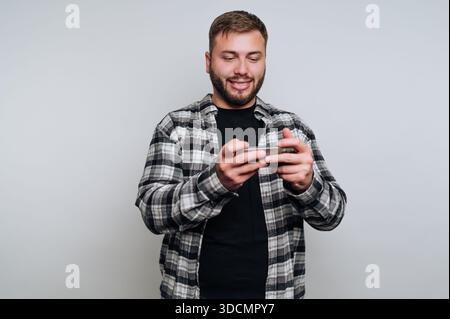 Ein junger Mann mit Bart lächelt und hält ein Smartphone in der Hand. Er steht vor einem einfachen Hintergrund, konzentriert sich auf seinen Bildschirm und engagiert sich in einem Stockfoto