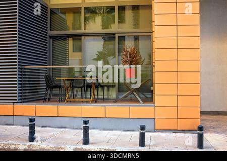 Stilvolle Loggia im Freien mit Holztisch und schwarzen Stühlen hinter Glasgeländern des modernen Gebäudes mit orangefarbenen Fliesen und Metallpaneelen. Stockfoto