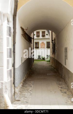 Ein bogenförmiger Steintunnel umrahmt den Blick auf ein weißes historisches Gebäude mit braunen Fensterläden und einem verblassten religiösen Fresko. Eine hölzerne Leiter hängt an Stockfoto