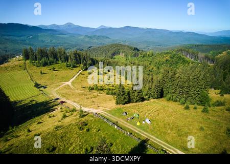 Campingplatz aus der Vogelperspektive in den Bergen, mit mehreren bunten Zelten auf einer grasbewachsenen Rodung. Camper entspannen unter klarem blauen Himmel, umgeben von hohen immergrünen und Panoramablick auf entfernte Gipfel. Stockfoto