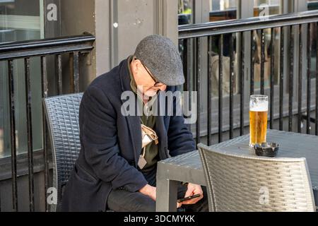 Ein älterer Mann mit flacher Mütze, der mit einem Bier an einem Tisch sitzt und auf sein Smartphone blickt. Stockfoto