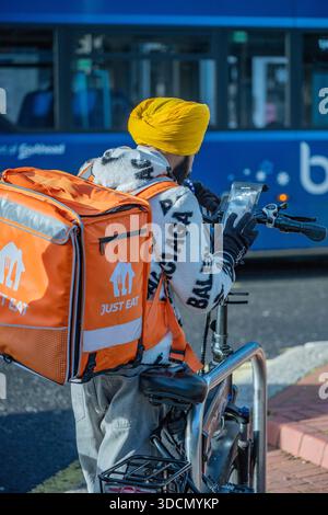 Essen Sie einfach einen Fahrradfahrer im Stadtzentrum von Southampton, Hampshire, Großbritannien. Essen Sie einfach einen Liefermann mit einem knallgelben Turban, der auf das Telefon schaut. Stockfoto