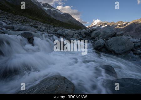 Eine Nahaufnahme aus der Nähe des schnellen, weißen Gletscherwassers, das über große graue Felsbrocken rauscht, mit dem sonnendurchfluteten, schneebedeckten Aoraki Mount Cook Peak i Stockfoto
