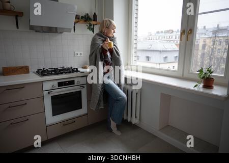 Frau mittleren Alters, die warm in kalten, unbeheizten Wohnungen bleibt, sich in kariertes Design einhüllt und heißen Tee trinkt. Stockfoto