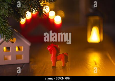 Rote dala-Pferdefigur auf einer Holzfläche, umgeben von leuchtendem Miniaturhaus, grünen Tannenzweigen und warmen Bokeh-Lichtern, die eine gemütliche Atmosphäre schaffen Stockfoto