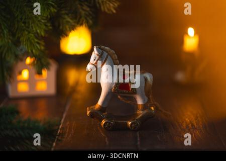 Miniaturspielzeug mit Schaukelpferd, das eine festliche weihnachtsszene auf einem Holztisch dekoriert, mit Keramikhaus, grünen Kiefernzweigen und warmer Atmosphäre Stockfoto