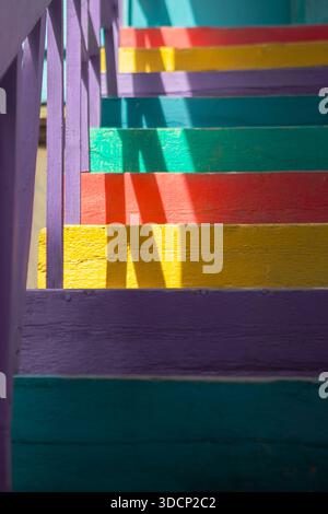 Regenbogenhölzerne Treppen mit violettem Geländer werfen scharfe diagonale Schatten über Stufen in hellem Sonnenlicht. Stockfoto
