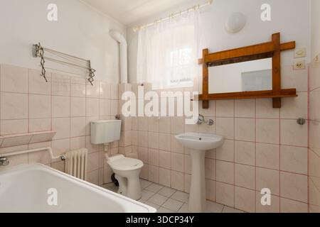 Die Innenausstattung des Badezimmers ist altmodisch und mit hellrosa Keramikfliesen Das Zimmer verfügt über eine weiße Badewanne, ein Sockelwaschbecken, eine Toilette und einen Spiegel mit Holzrahmen. Alt Stockfoto