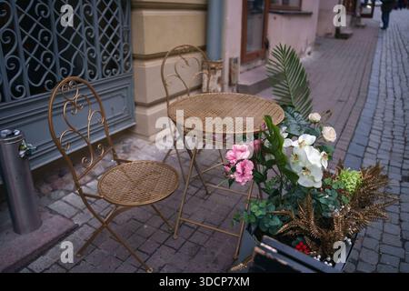 Charmantes Straßencafé mit kunstvollen Metallmöbeln und lebhaften Blumenarrangements vor einem Kopfsteinpflasterweg in einer malerischen urbanen Gasse, Design Stockfoto