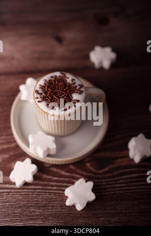 Tasse cremige heiße Schokolade mit Kakaostreuseln auf einem Holztisch, umgeben von sternförmigen Marshmallow-Leckereien, die an eine warme und gemütliche Winte erinnern Stockfoto