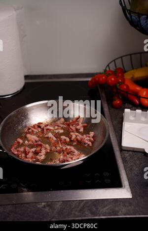 Brutzelnde Speckstreifen, die in einer Edelstahlpfanne auf Induktionsherd gekocht werden, umgeben von frischen Produkten wie Tomaten und Bananen in der modernen Küche Stockfoto