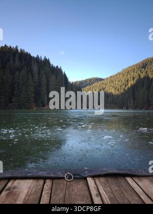 Roter See Rumänien, gefrorener Bergsee umgeben von dichtem Kiefernwald unter klarem Himmel, Blick von einem Holzpier in einer friedlichen Winterlandschaft. Stockfoto