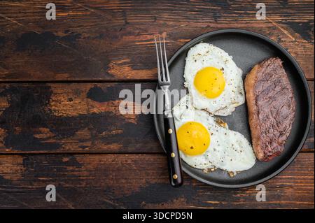 Traditionelles Steak und Eier Frühstück mit gebratenem Rindfleisch und sonnigen Eiern, serviert in gusseiserner Pfanne, Blick von oben auf Holztisch. Stockfoto