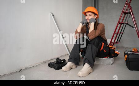 Nachdenkliche Reparaturfrau, Vorarbeiter oder Baumeister in Uniform und Schutzhelm, die in unvollendetem Gebäude sitzt. Reparatur, Bau und Gebäudekonzept. Banner Stockfoto