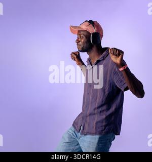 Junger schwarzer Mann, der Kopfhörer trägt und im Studio Musik genießt, entspanntes Lifestyle-Konzept mit violettem Hintergrund und Kopierraum Stockfoto