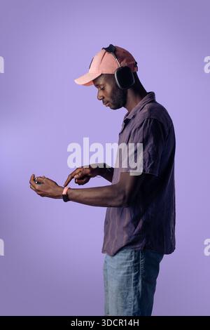 Junger schwarzer Mann, der Kopfhörer trägt und ein Smartphone in einer Studioumgebung benutzt, Seitenprofil mit violettem Hintergrund und Kopierraum, modernes Lif Stockfoto