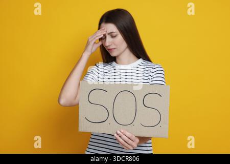 Besorgte Frau, die ein Schild mit dem Wort SOS auf gelbem Hintergrund hält Stockfoto