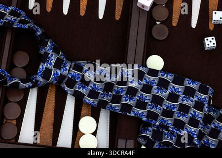 Nahaufnahme einer bunten Krawatte aus Seide in Schwarz, Blau und Silber, die über Backgammon-Stücke und Würfel hängt und elegante Mode mit Freizeit verbindet Stockfoto