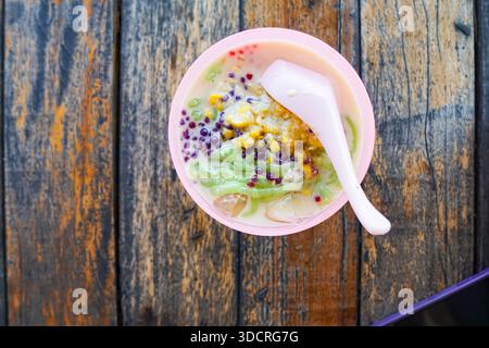 Cendol, ein malaysisches Dessert, das in einer roten Schüssel mit zerstoßenem Eis, grünem Gelee, roten Perlen, Kokosmilch und klebrigem Reis auf einem rustikalen Holztisch serviert wird. Stockfoto