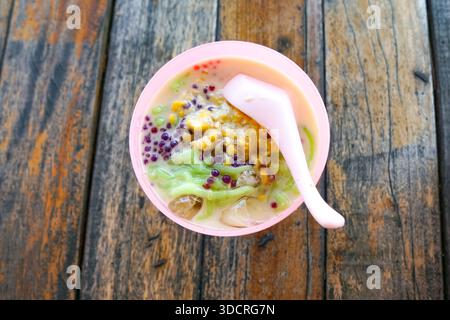 Cendol, ein malaysisches Dessert, das in einer roten Schüssel mit zerstoßenem Eis, grünem Gelee, roten Perlen, Kokosmilch und klebrigem Reis auf einem rustikalen Holztisch serviert wird. Stockfoto